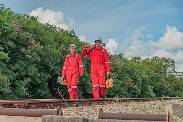 Twee medewerkers in rode duurzame werkkleding met reflecterende elementen werken buiten langs het spoor.