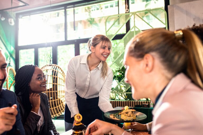 Serveerstermedewerkster in stijlvolle hospitality kleding bedient lachend gasten in een modern restaurant.